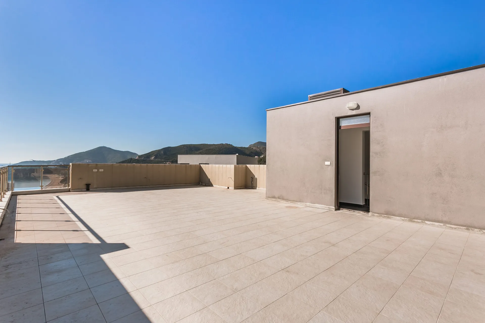 Large open tiled terrace with glass railing overlooking mountains and sea under a clear blue sky.