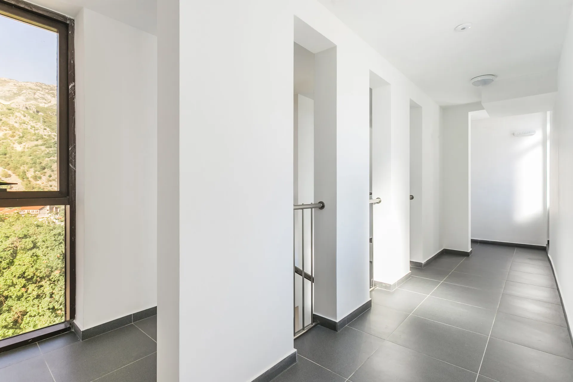 Long white hallway with grey tile flooring, a metal stair railing, and a window overlooking hills.