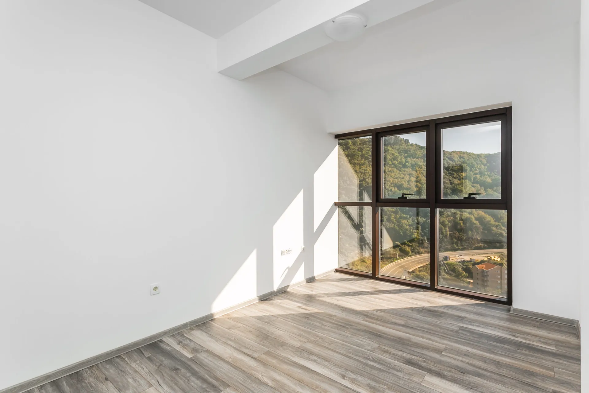 Empty room with white walls, grey laminate flooring, and a large window overlooking a forested hillside.