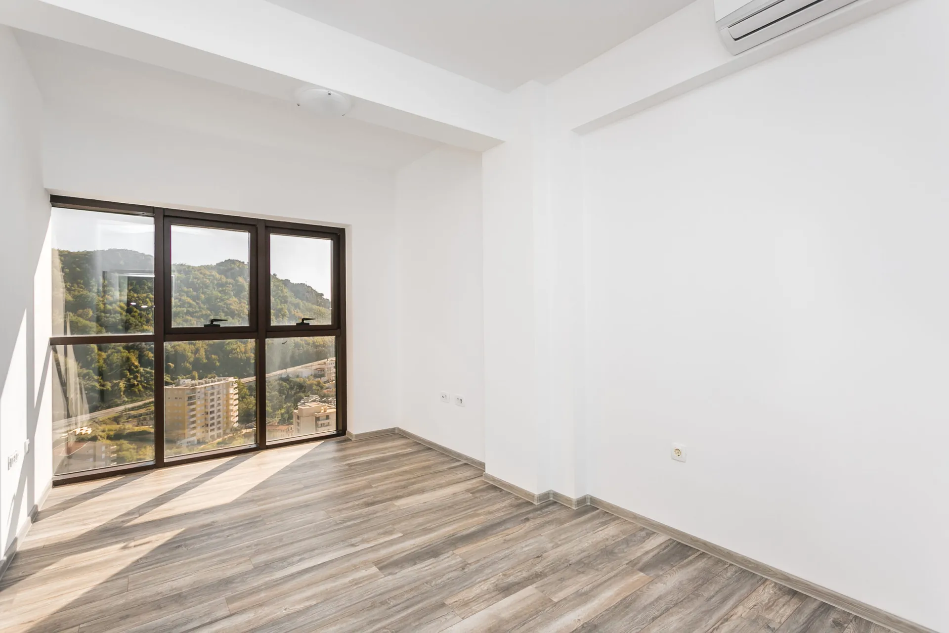 Empty room with laminate flooring and large window overlooking forested hills and city buildings.