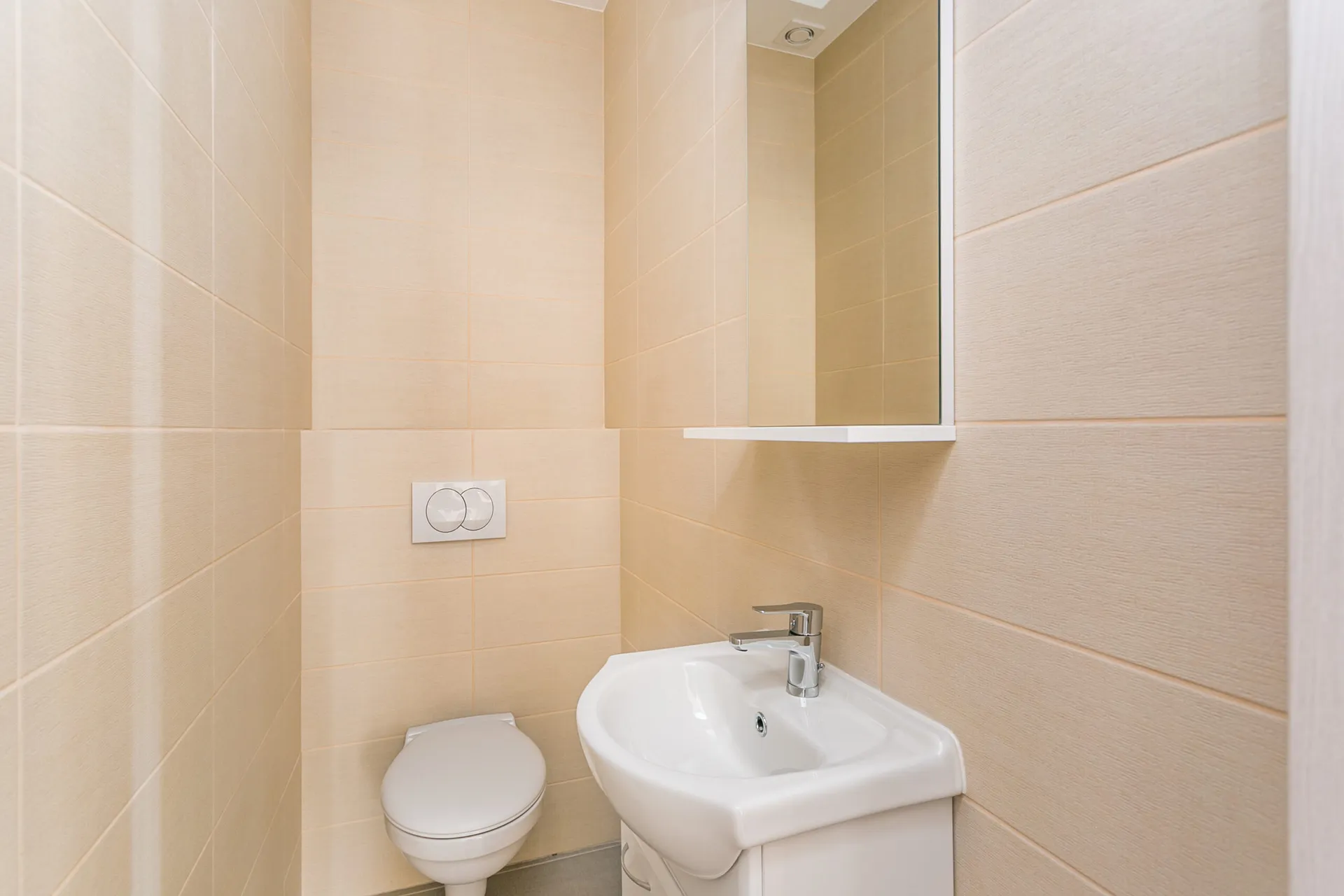 Small bathroom with beige tiles, white toilet, sink, and wall mirror featuring a chrome faucet.