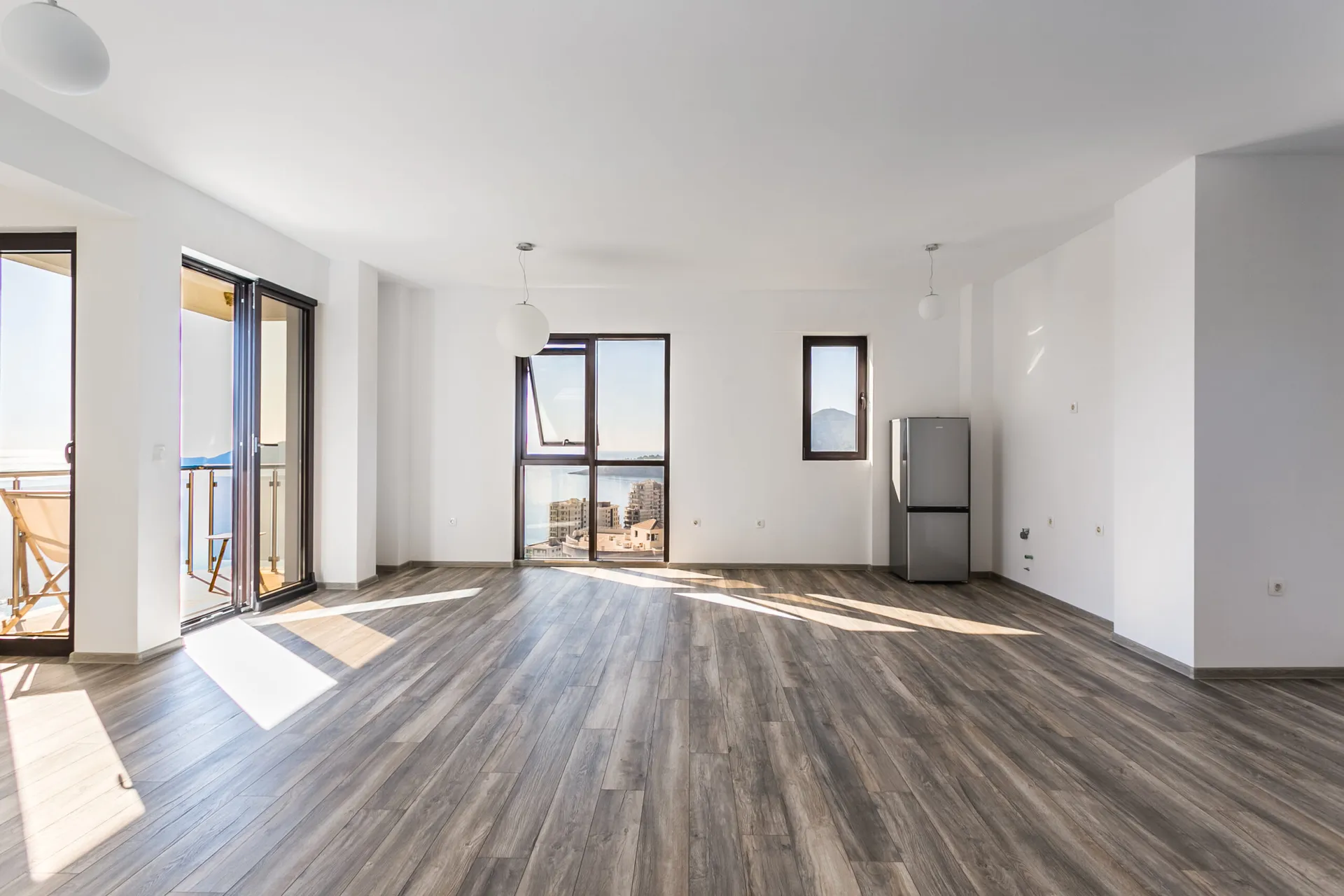 Empty room with wood flooring, a refrigerator, and balcony doors overlooking the sea.