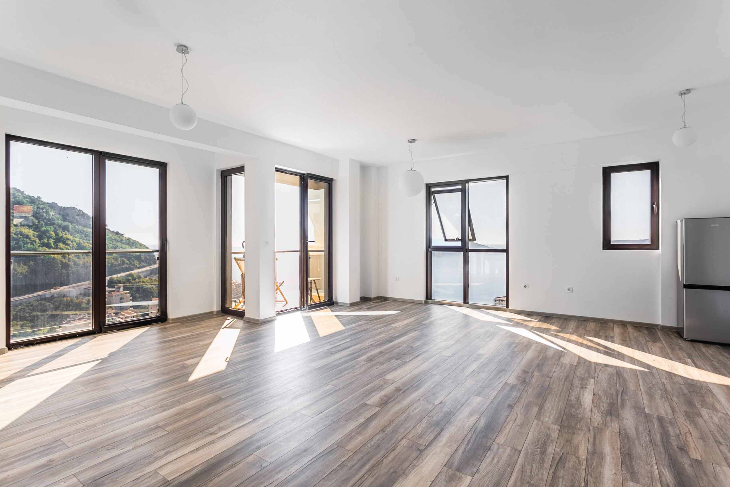Bright living room in a penthouse in Rafailovici, Montenegro, with panoramic windows, sea and mountain views, and wood-look flooring.