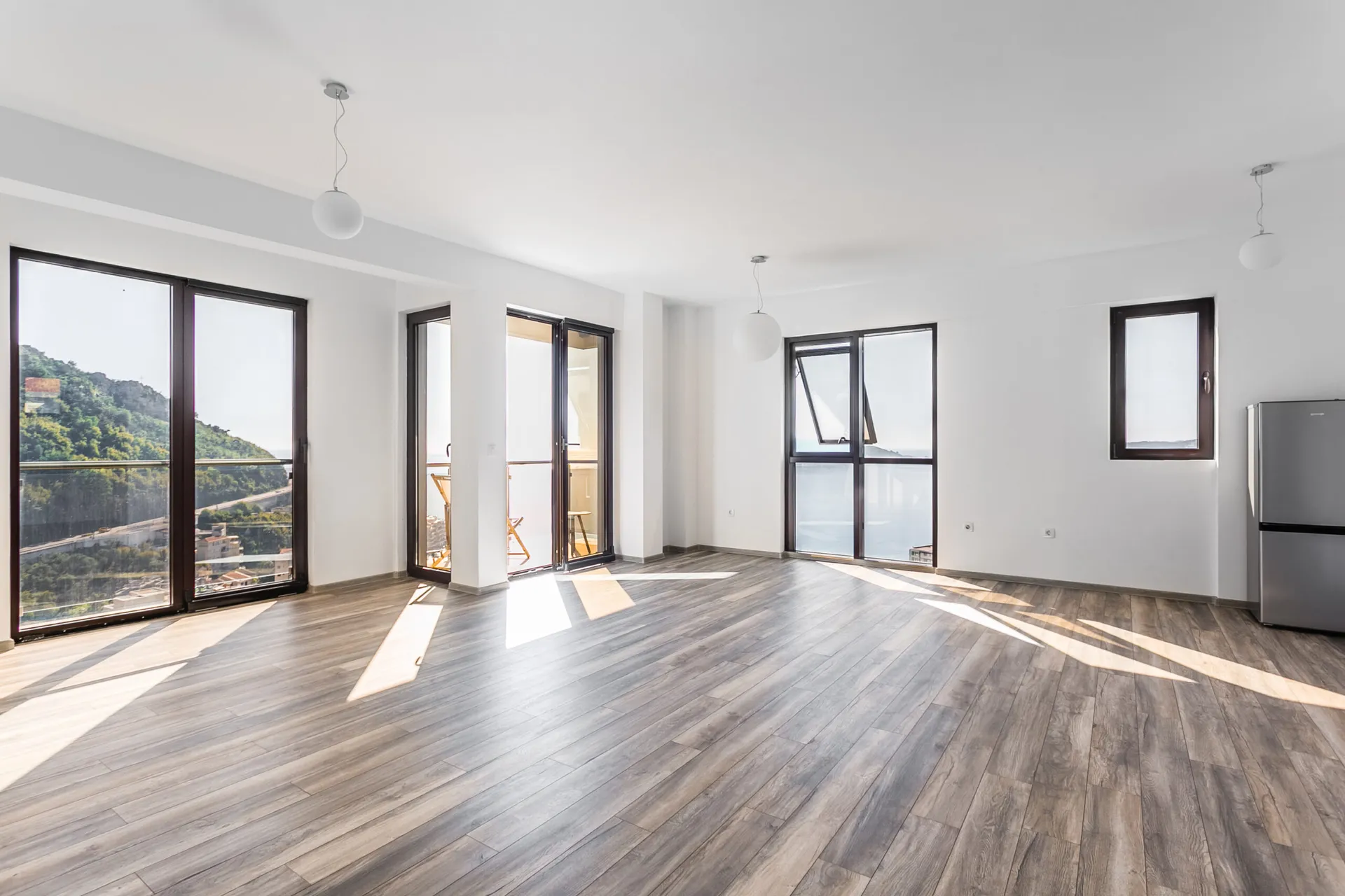 Bright empty room with wood flooring, large windows, balcony access, and a refrigerator on the right.