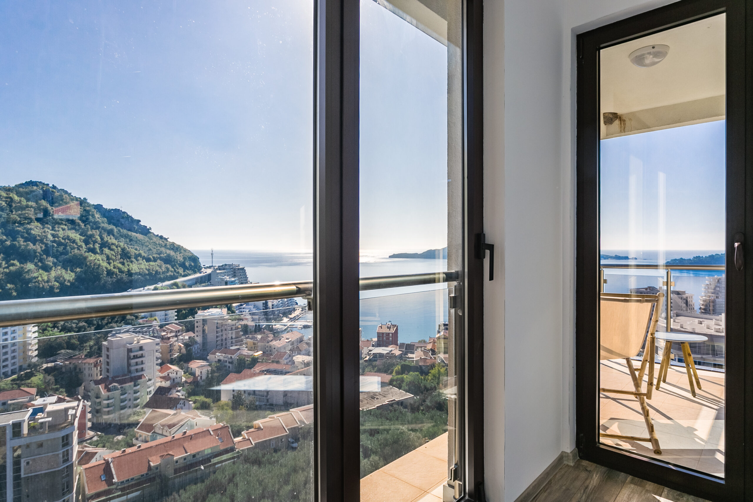 View from the penthouse balcony in Rafailovici, Montenegro: a panorama of the sea, mountains, and city through a glass railing.
