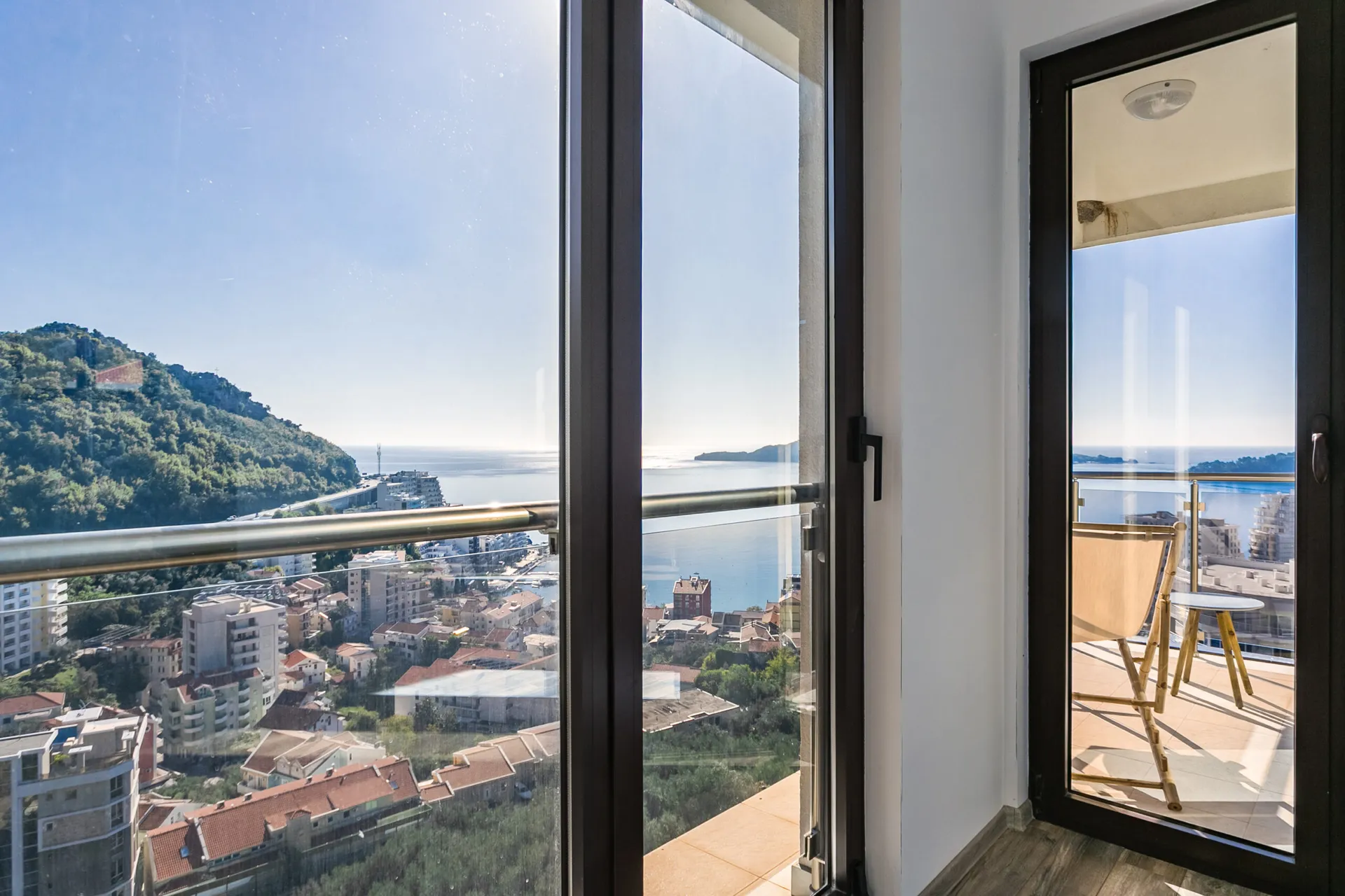 View from an interior window onto a balcony with a chair and table overlooking the city and sea.