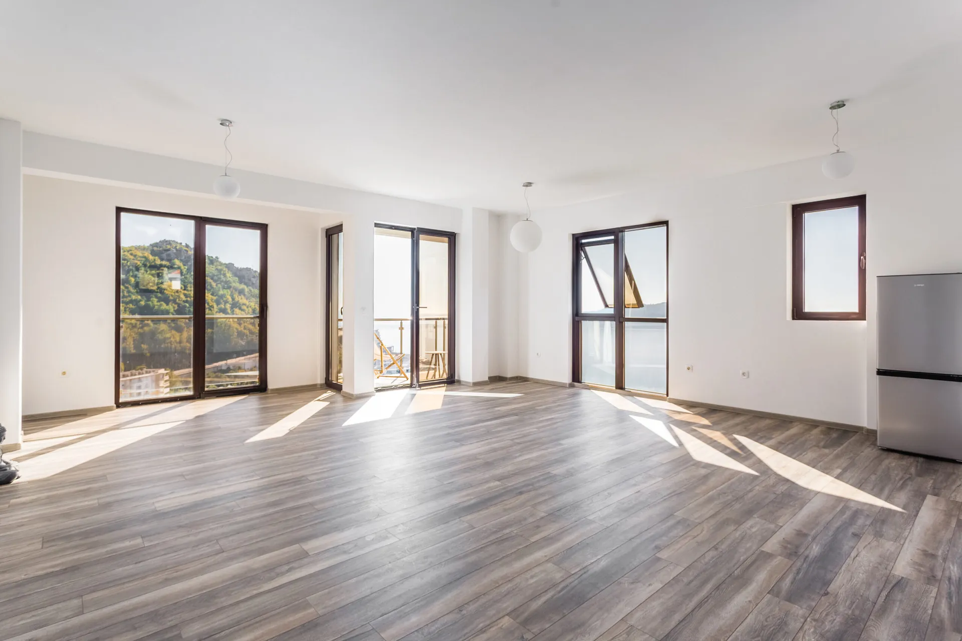 Empty room with wood laminate flooring, large windows, balcony access, and a refrigerator on the right.