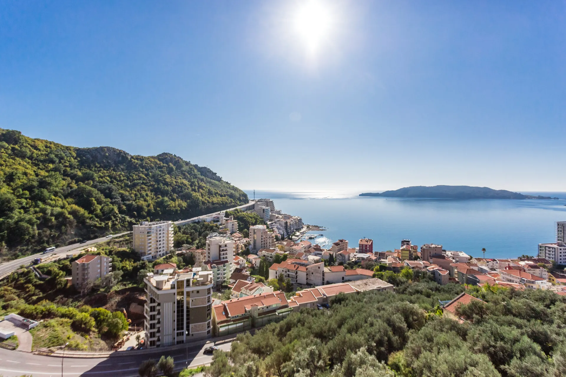 Panoramic exterior view of a coastal town, sea, and green hill under bright sun.