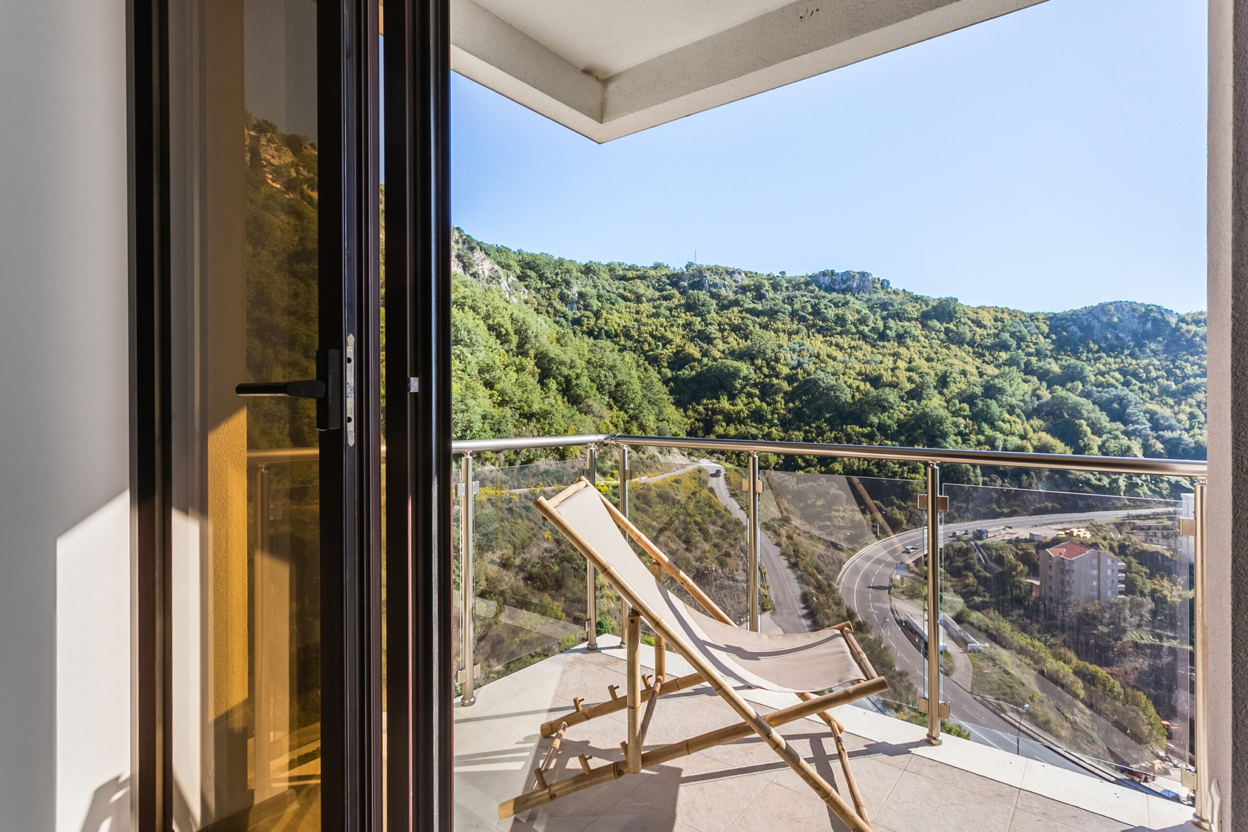 Penthouse balcony in Rafailovići, Montenegro: a sun lounger, glass railing, and panoramic view of the mountains and road.
