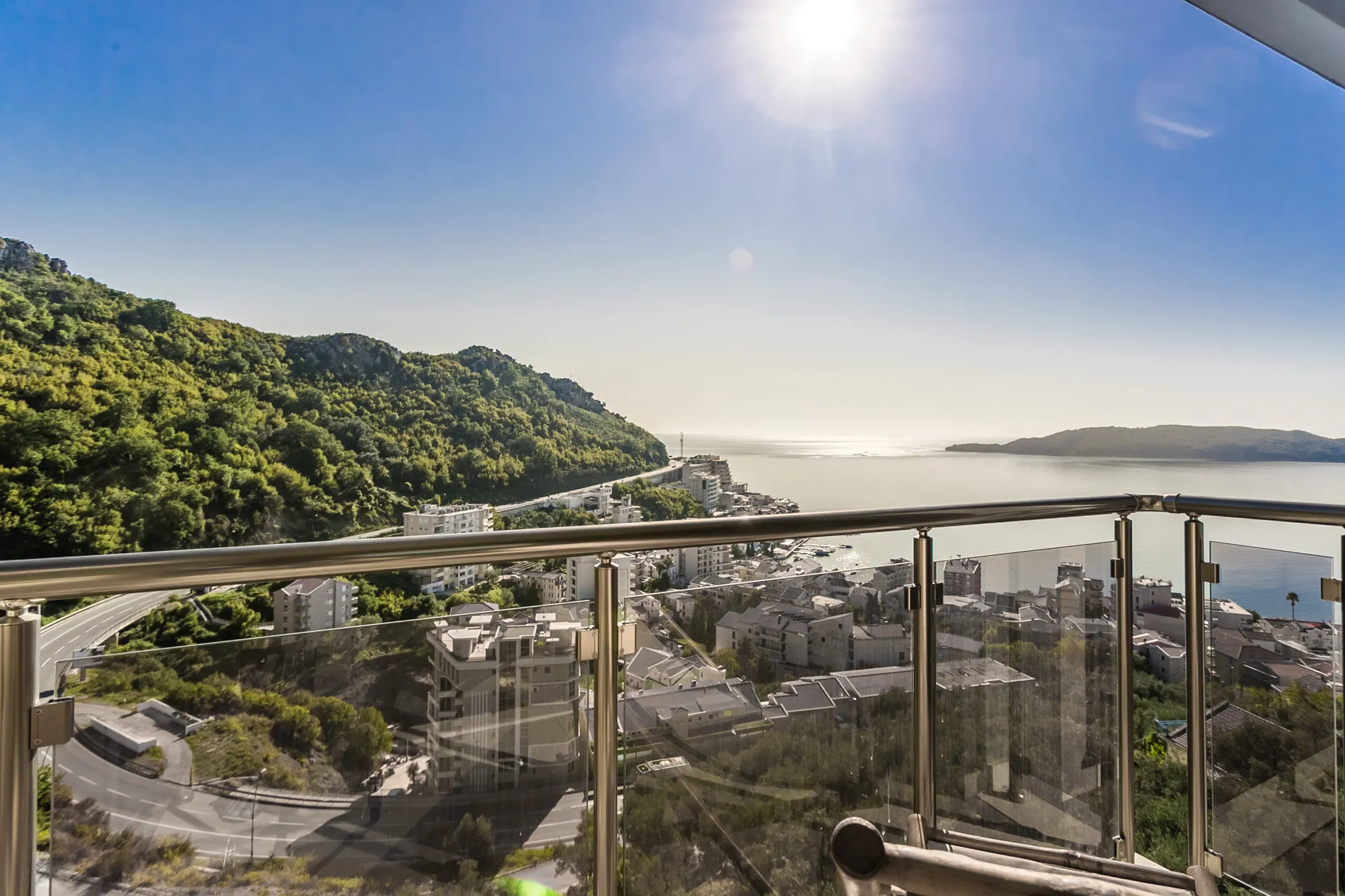View from a balcony with glass railing overlooking the sea, a green hill, and coastal town under bright sun.