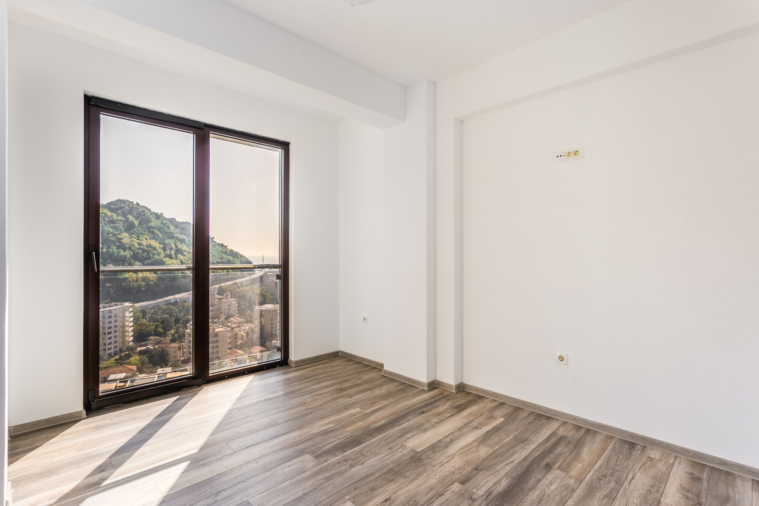 Bright living room in a penthouse in Rafailovici, Montenegro: panoramic window with mountain views, laminate flooring, and white walls.