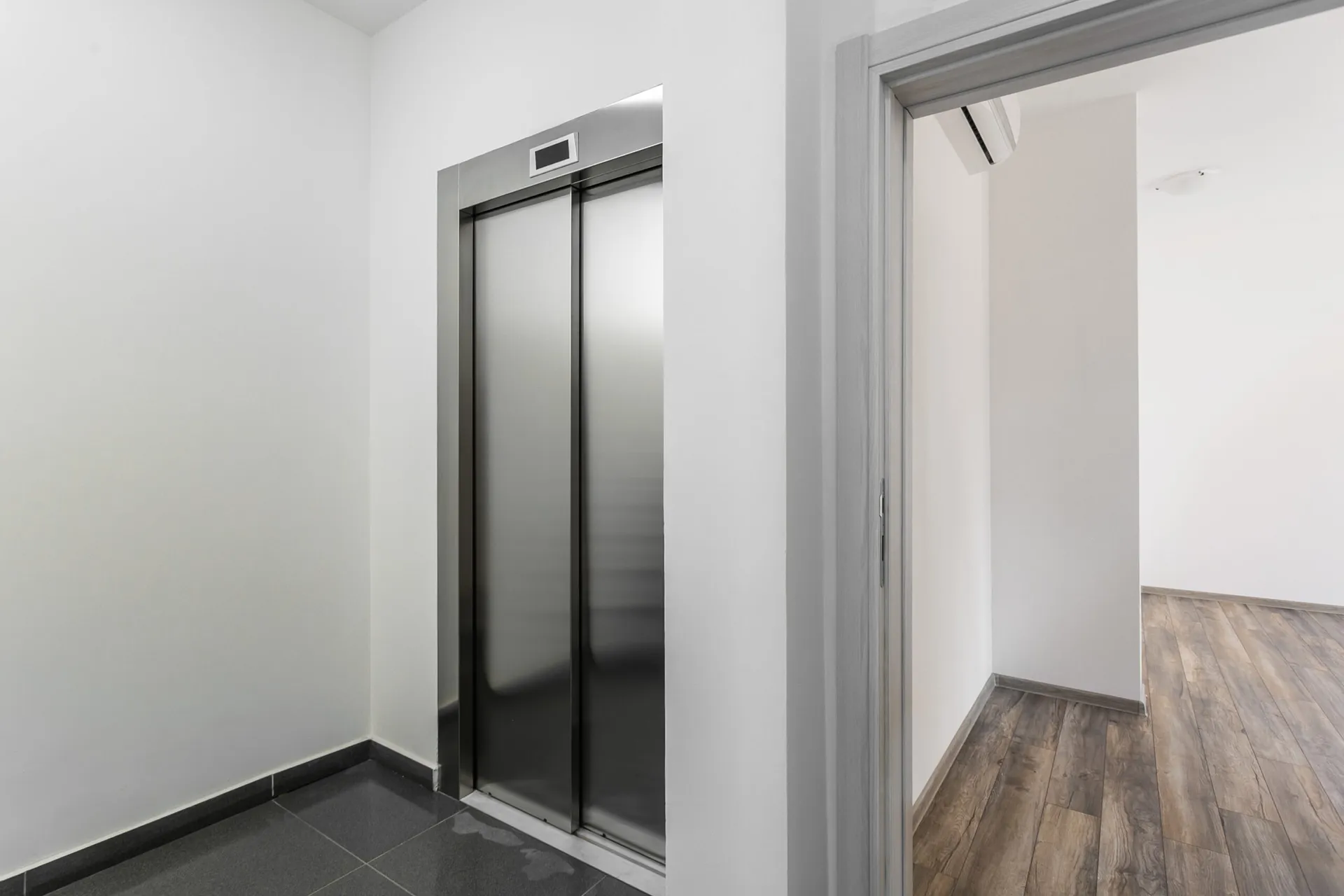 Elevator lobby with stainless steel doors and grey tile flooring, adjacent doorway leads to a room with wood laminate flooring.