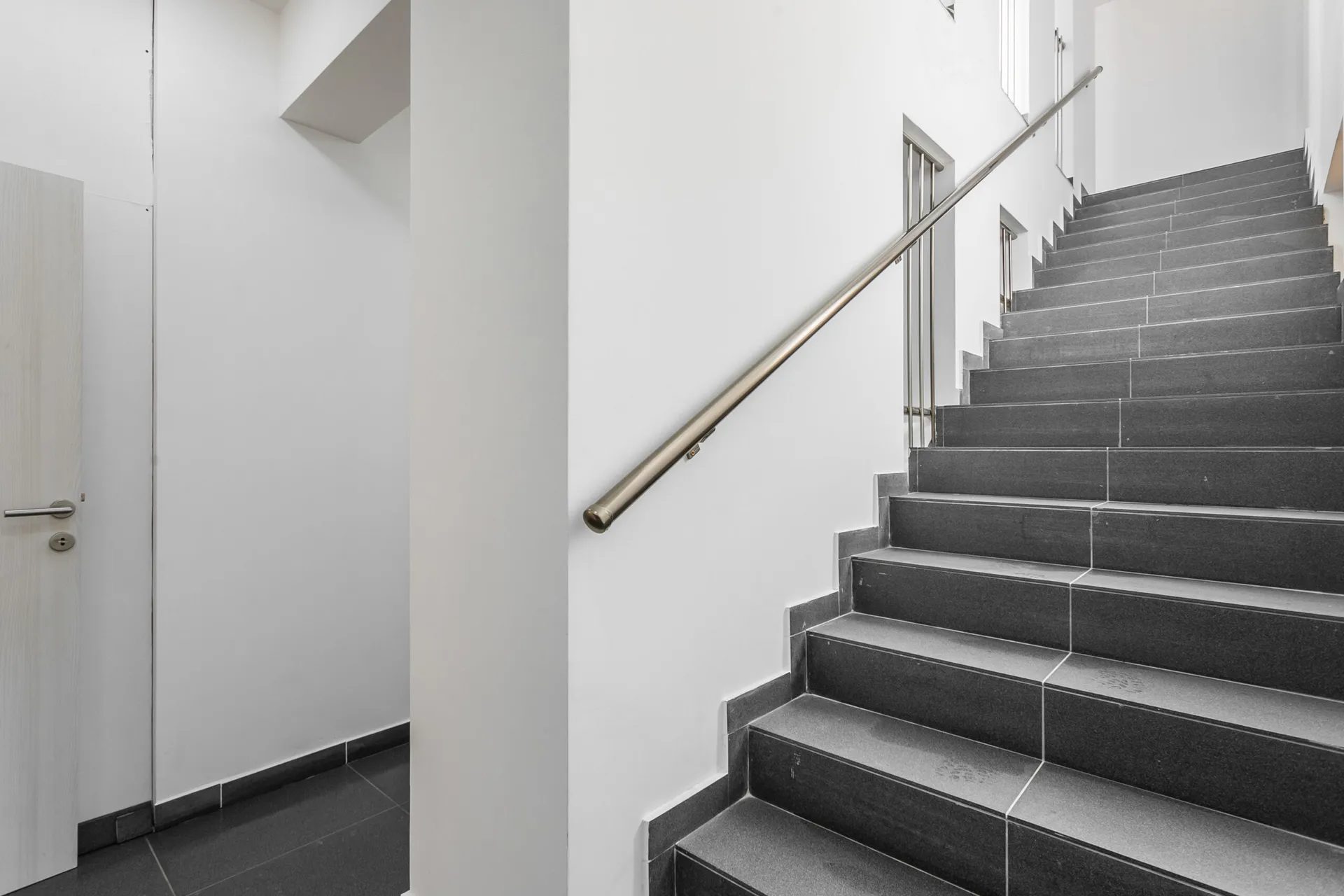 Staircase with dark tiles and metal handrail on white wall next to a light wood door.