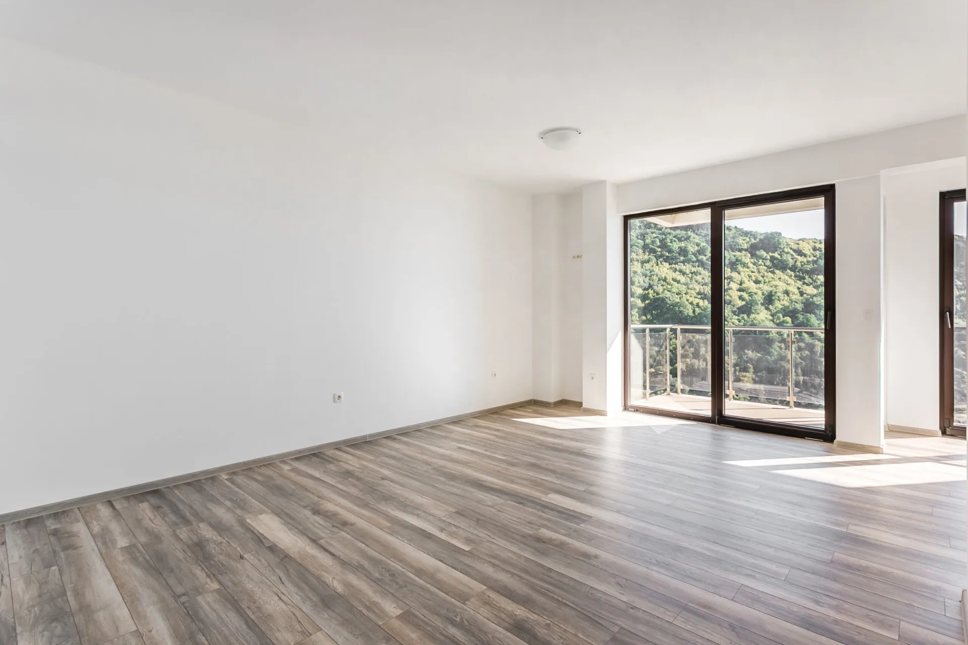 Empty room with grey laminate flooring and white walls. Large glass door leads to a balcony overlooking a forest.