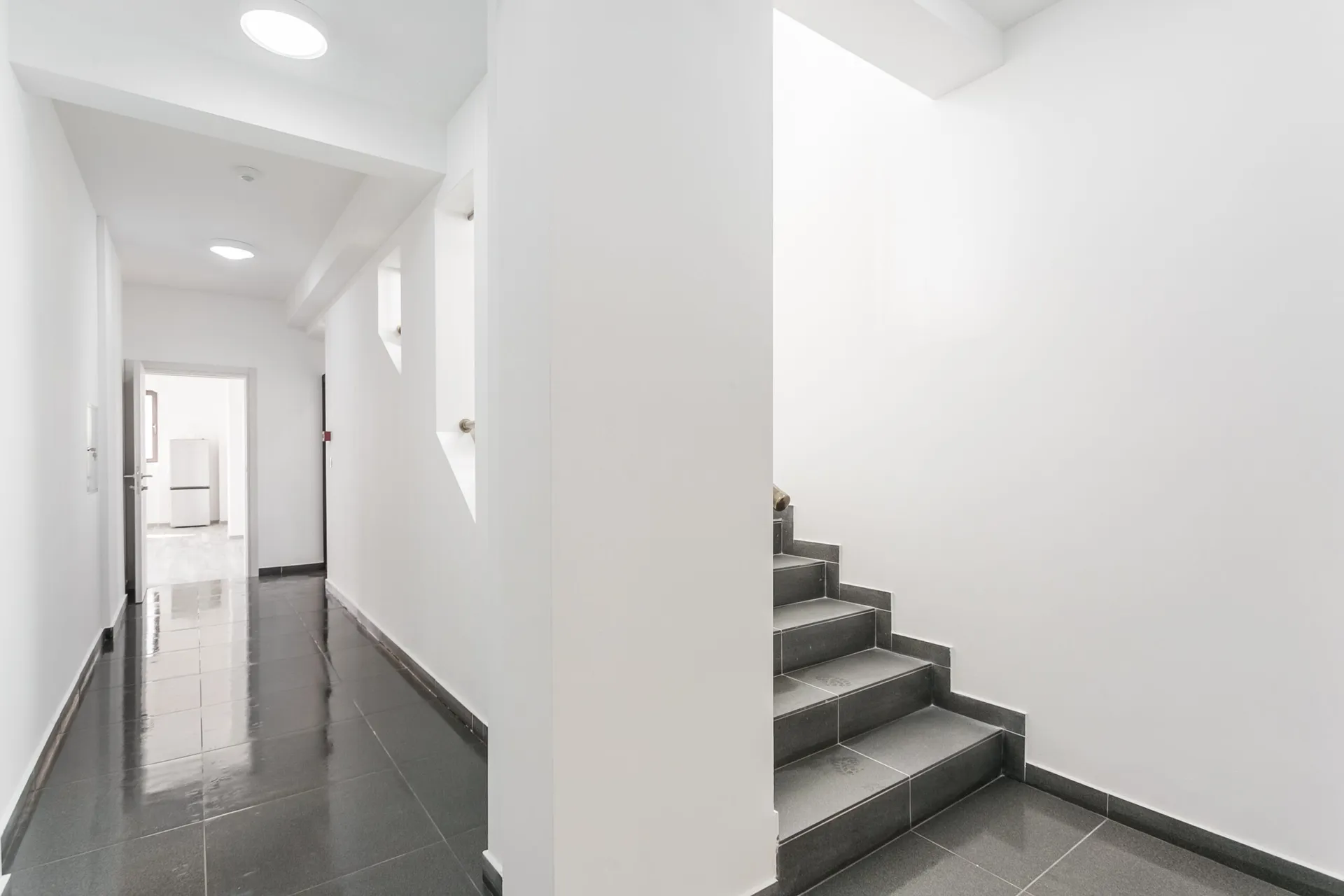 White hallway with dark tiled floor and staircase leading up, door visible at end.