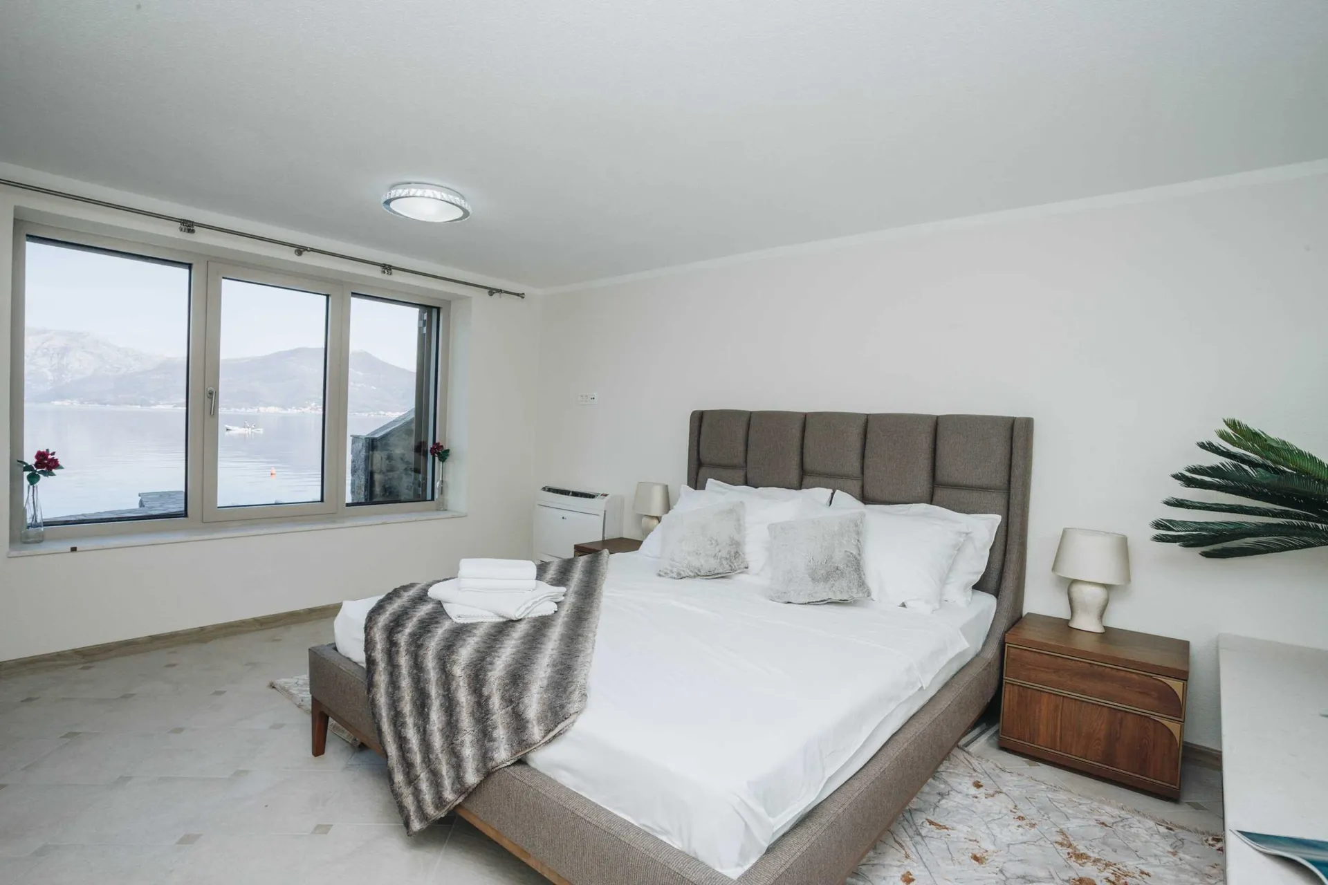 Bright bedroom with a double bed, wooden nightstand, and a large window overlooking the water.