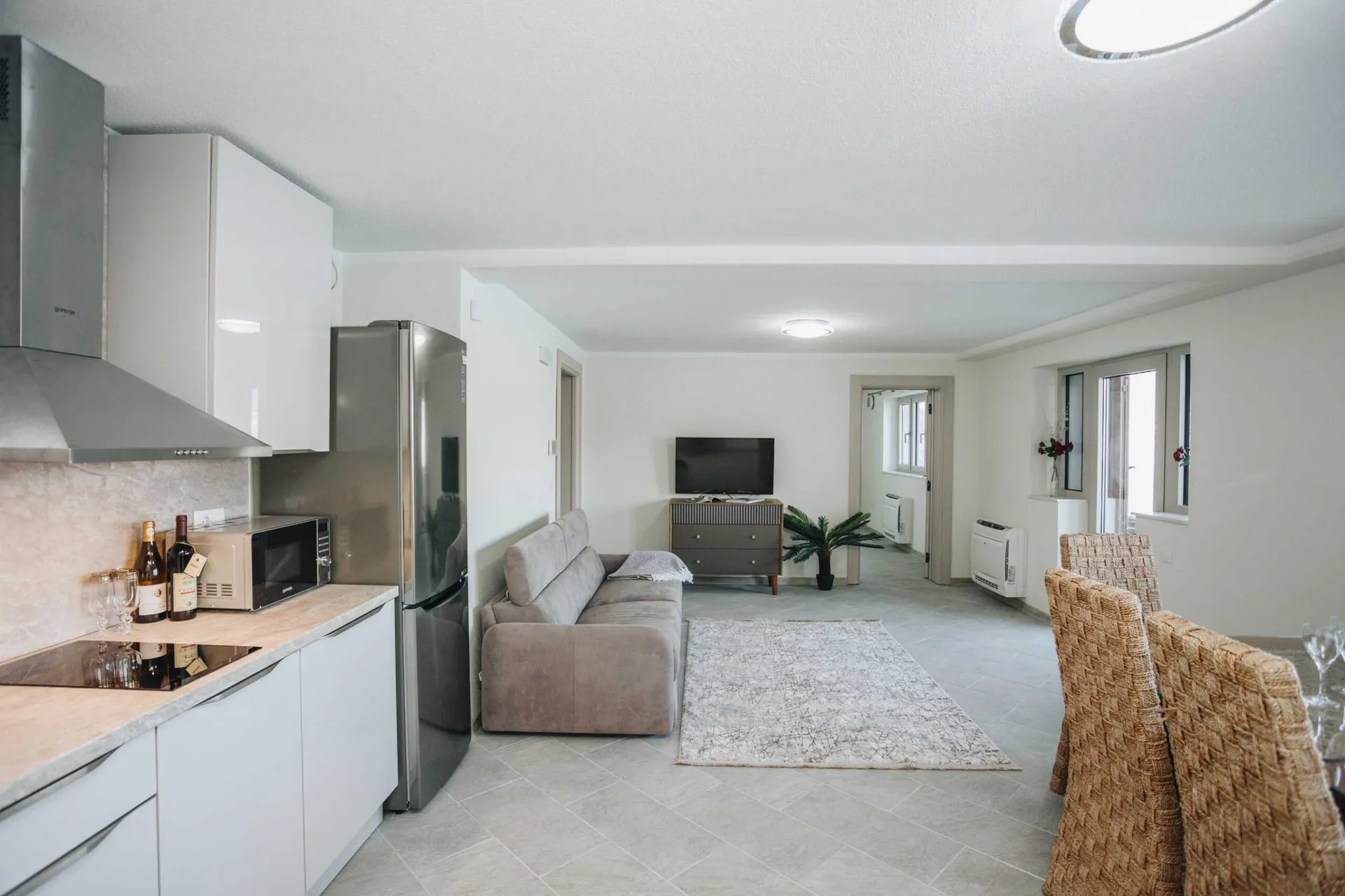 Open plan space featuring a kitchen area, grey sofa, TV unit, and woven dining chairs.