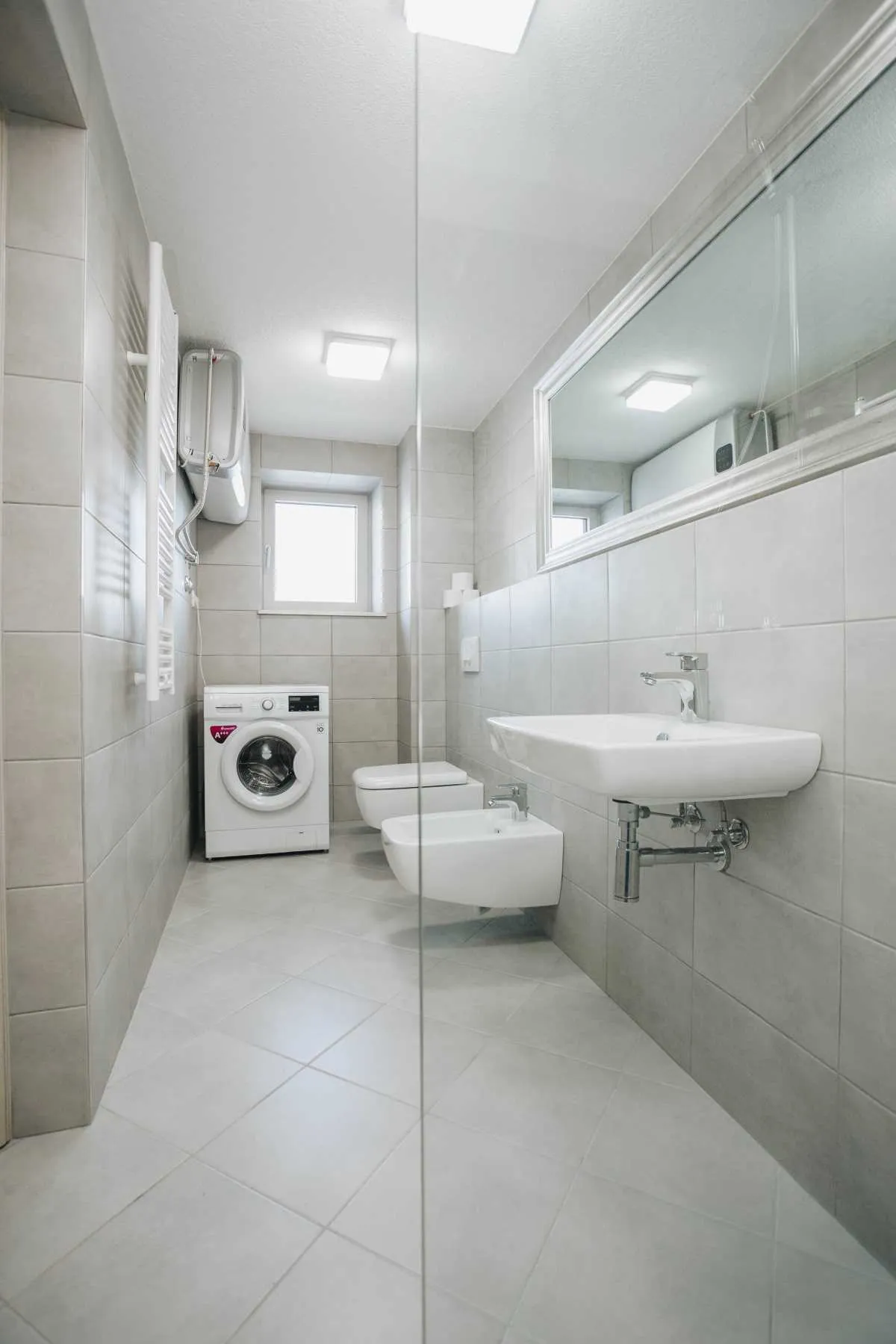 Modern bathroom with grey tiles, washing machine, toilet, bidet, and wall-mounted sink under a large mirror.
