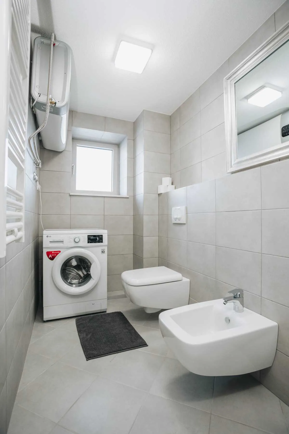 Bathroom with grey tiles, wall-mounted toilet, bidet, washing machine, water heater, and a small window.