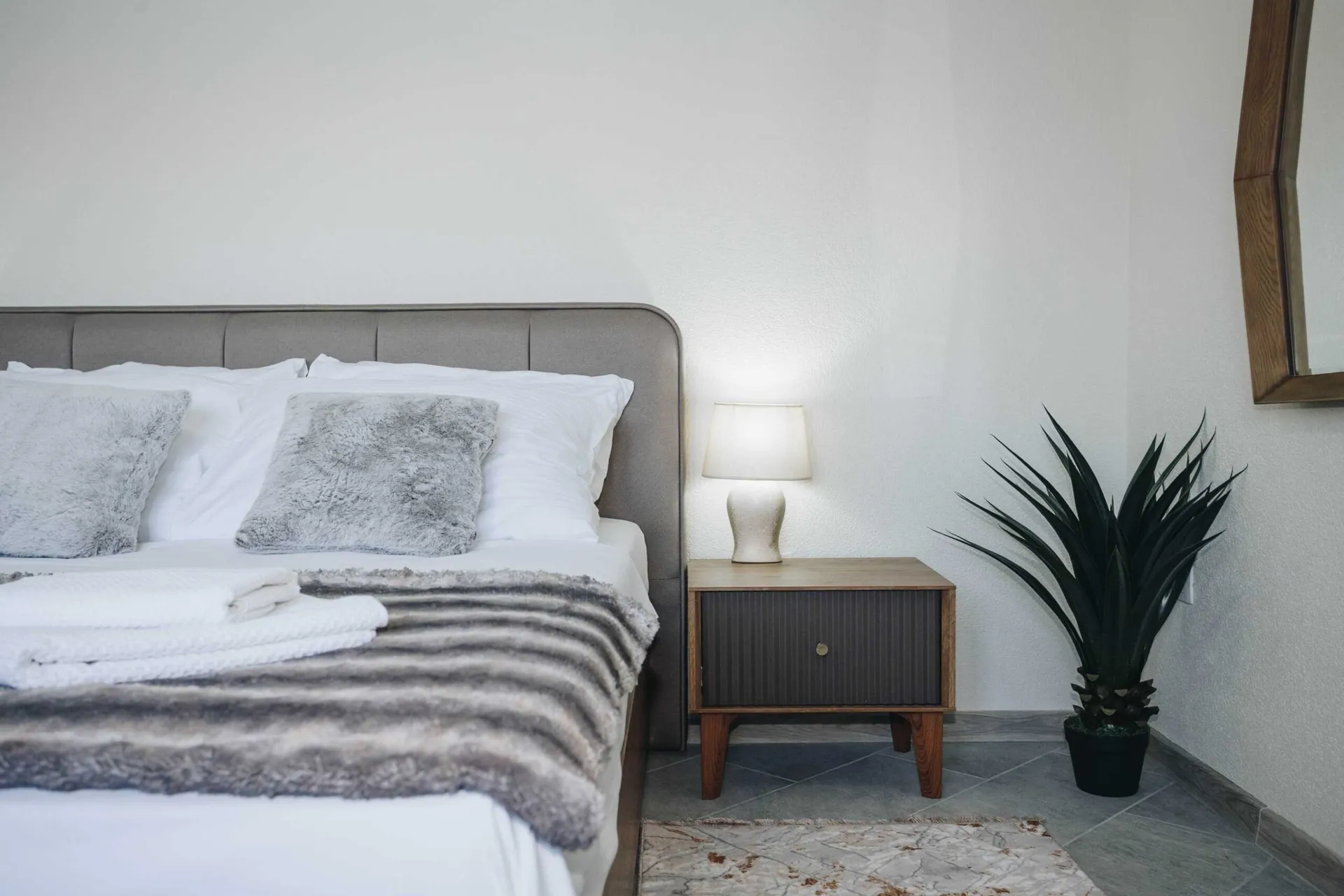 Modern bedroom featuring a grey upholstered bed, white bedding, a wooden nightstand with a lamp, and a potted plant.