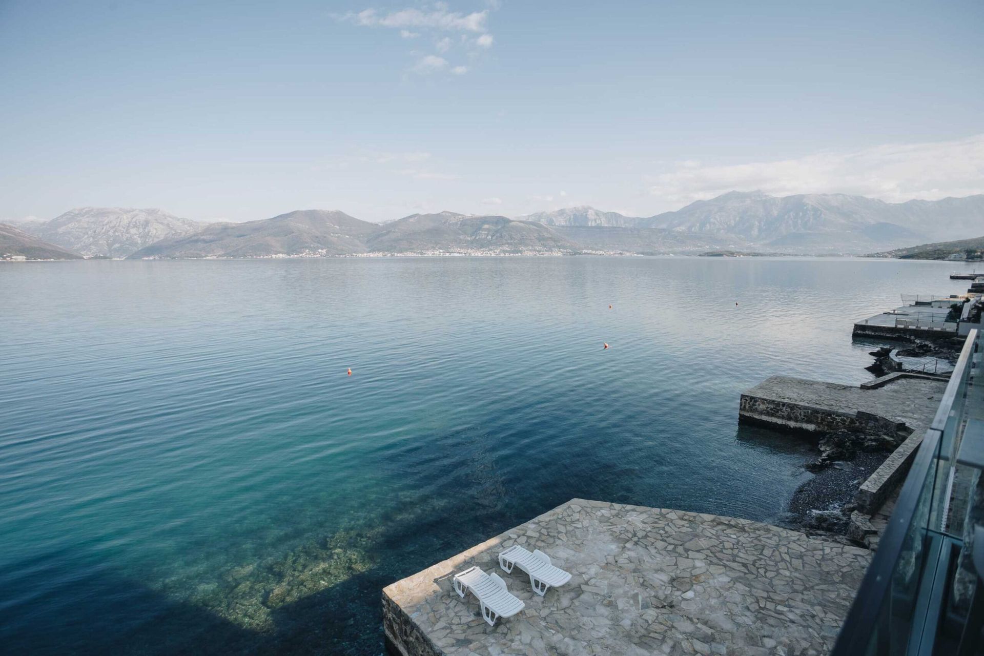Sea and mountain view from the villa’s private stone beach in Krašići, Montenegro, with white loungers by the water.