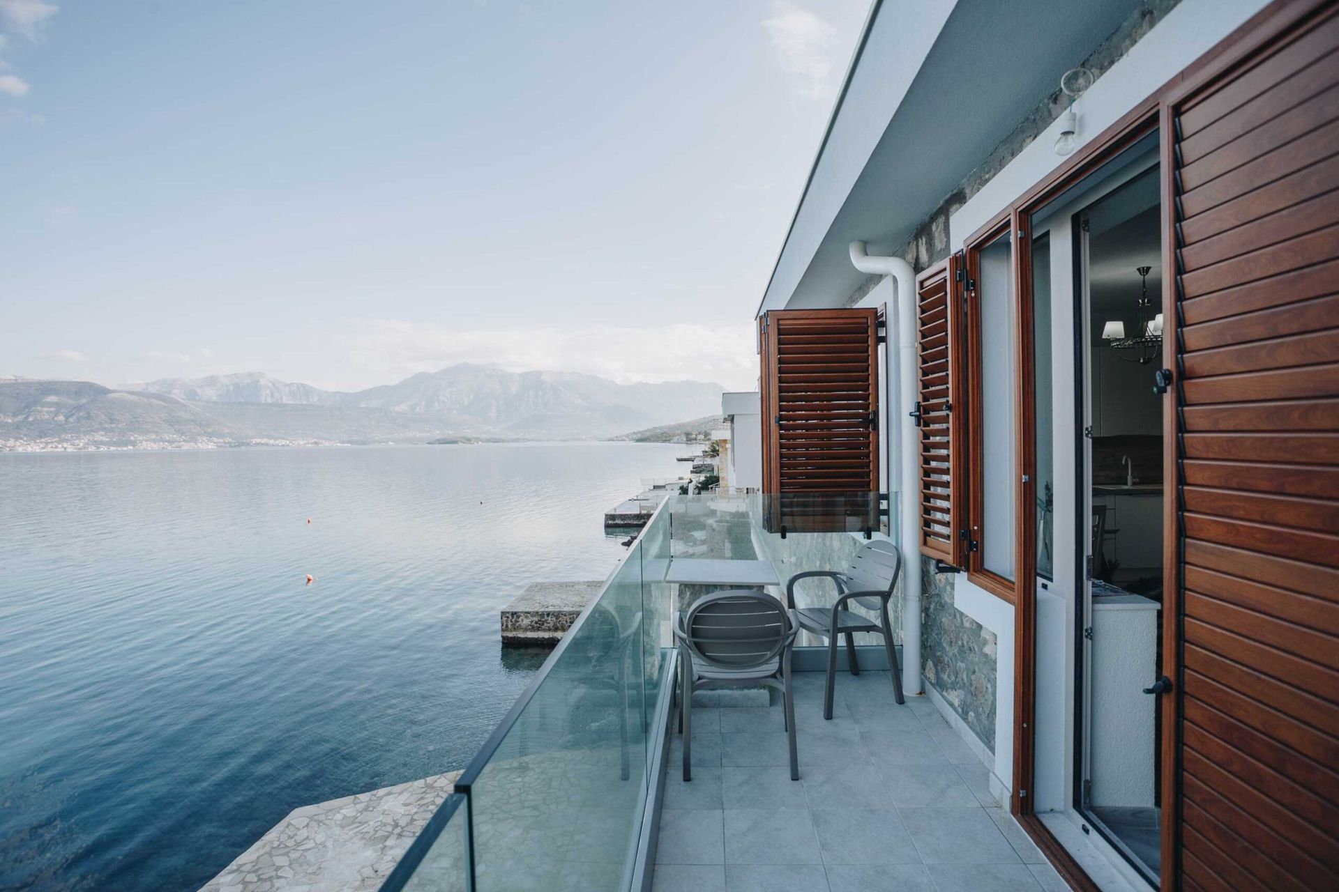 Villa balcony in Krašići, Montenegro: sea and mountain views, glass railing, wooden shutters, and lounge furniture.