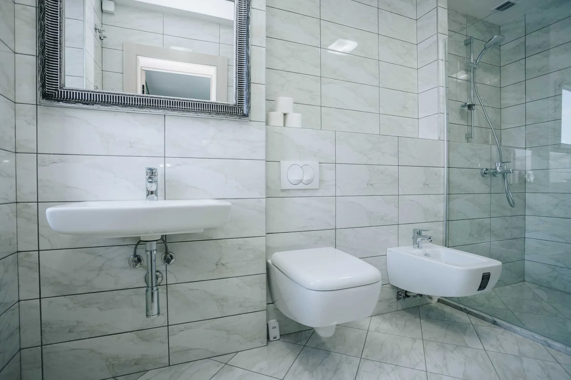 Modern bathroom with white marble tiles, wall-mounted toilet, bidet, sink, and glass-enclosed shower area.