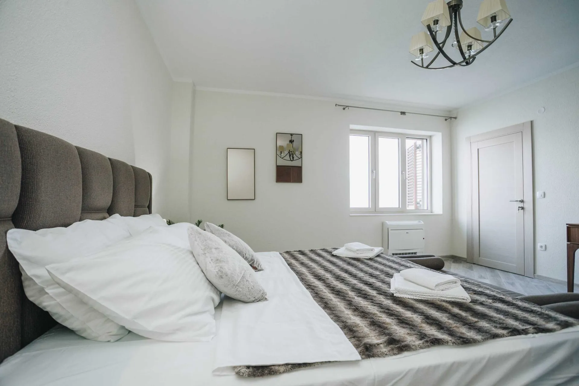Bright bedroom featuring a large bed with a grey upholstered headboard, white linens, and a patterned throw blanket.