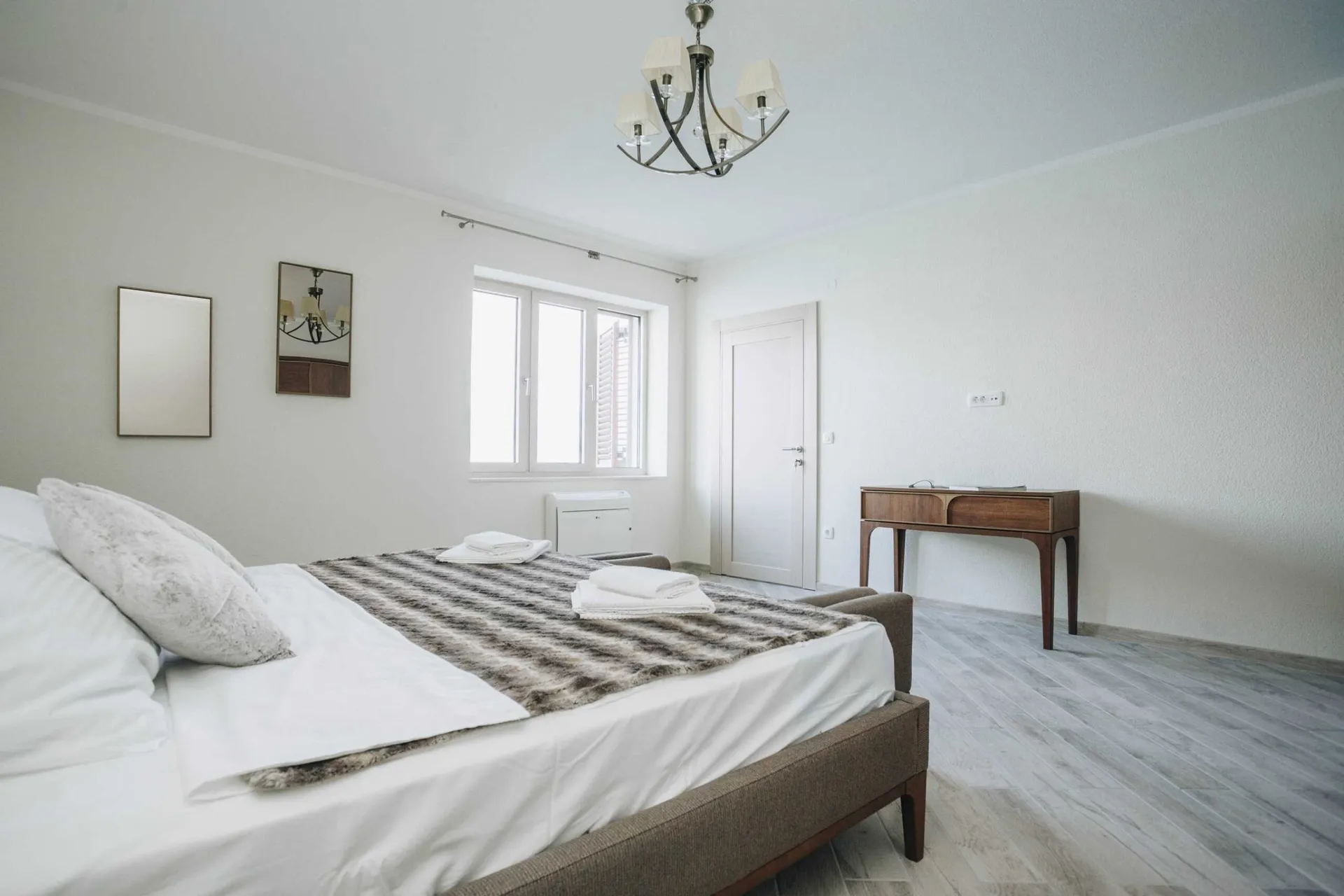 Bright bedroom featuring a double bed with towels, a wooden console table, chandelier, and wall mirror.