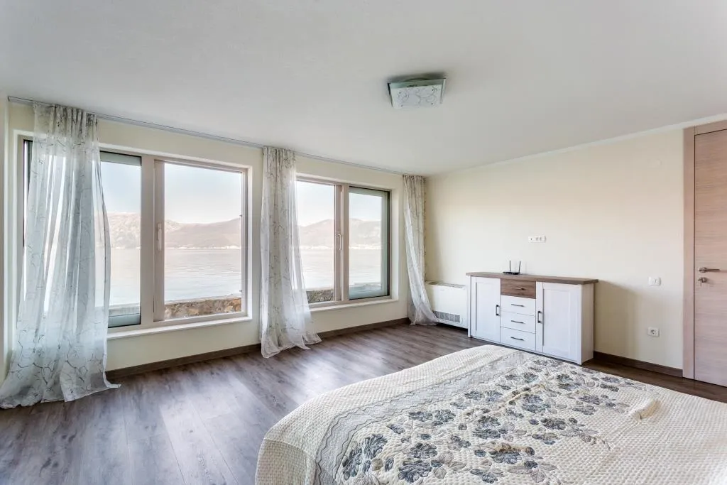 Bright bedroom featuring a large bed with floral coverlet, white dresser, and windows overlooking water and mountains.