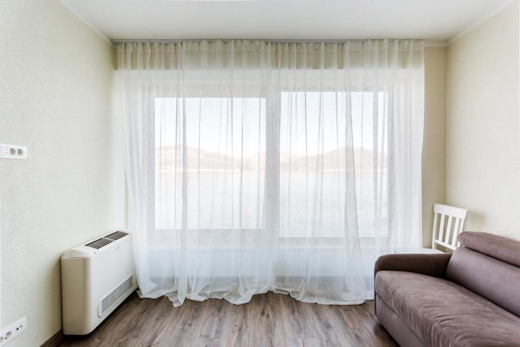Living room with sea view in a villa, Krašići, Montenegro: brown sofa, light walls, and a panoramic window with sheer curtains.