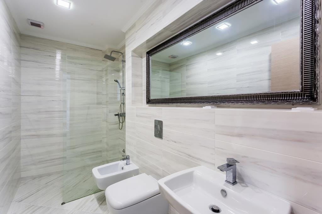 Modern bathroom with marble tiles, glass shower enclosure, toilet, bidet, sink, and a large framed mirror.