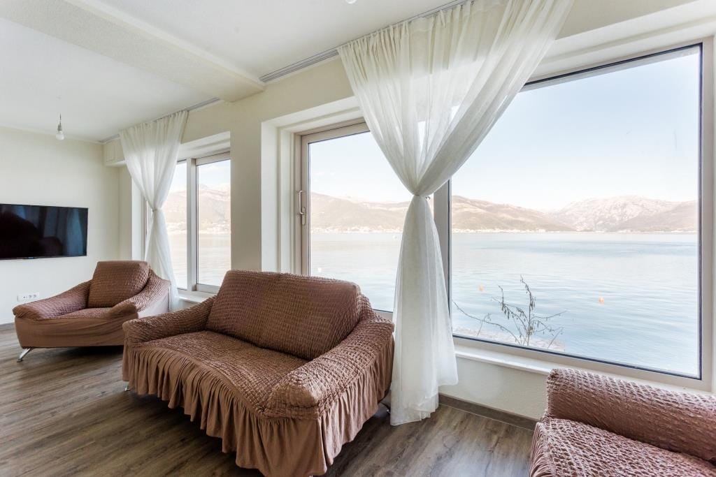 Living room with brown sofas, TV, and large windows overlooking water and mountains.