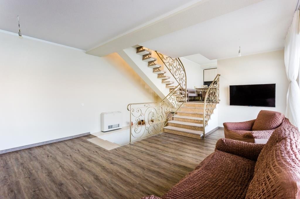 Living room with brown sofa, wall TV, and staircase featuring ornate metal railing on wood floor.