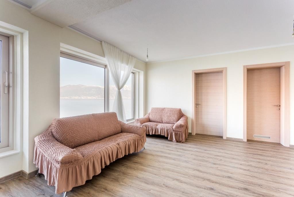 Bright living room in a villa in Krašići, Montenegro: two sofas, panoramic windows with sea and mountain views, light laminate flooring.