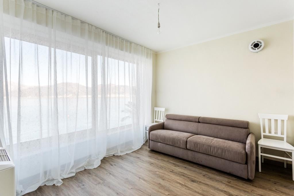 Living room with a brown sofa, white chairs, large window with sheer curtains overlooking water.