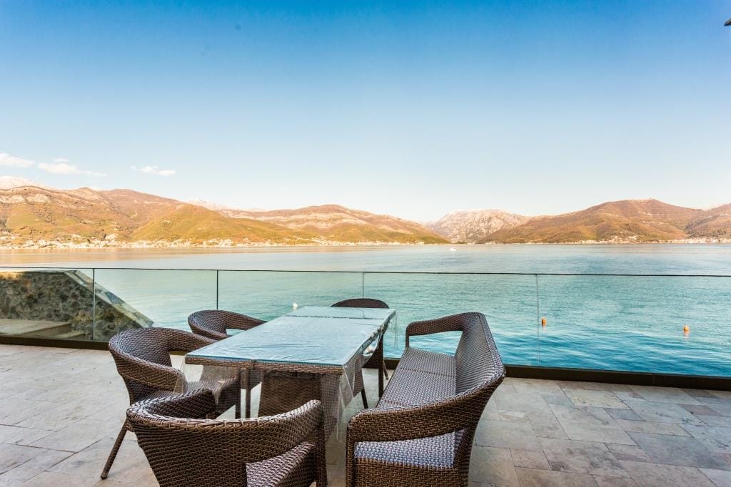 Terrace with wicker furniture and glass railing overlooking a lake and mountains.