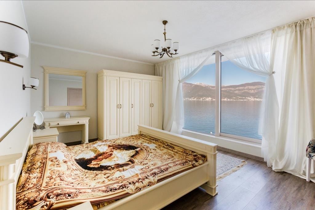Bright bedroom with a white bed, wardrobe, and vanity table. A large window offers a scenic view of the lake and mountains.