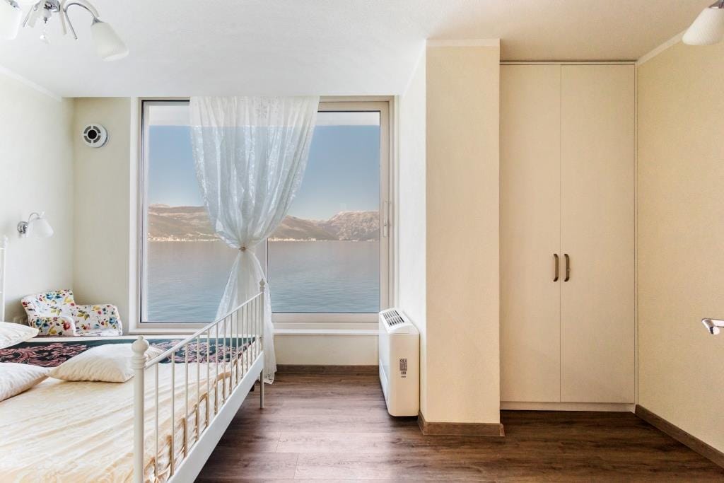 Bedroom with sea view in a villa, Krašići, Montenegro: white bed, built-in wardrobe, and panoramic window overlooking the bay.