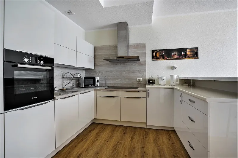 Modern kitchen with glossy white cabinets, built-in black oven, stainless steel hood, and wood-effect flooring.