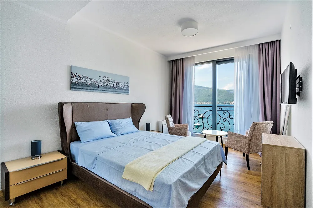 Bedroom with a double bed, blue bedding, and a balcony offering a scenic view of the sea and mountains.