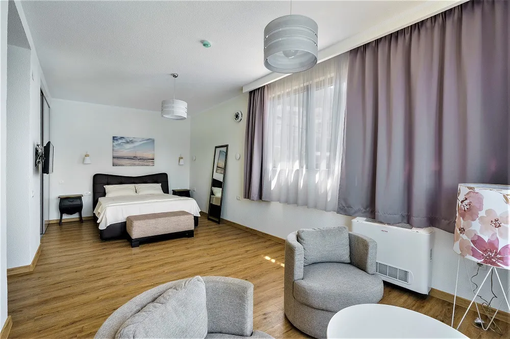 Bedroom featuring a double bed, grey armchairs, wooden flooring, and a large window with curtains.