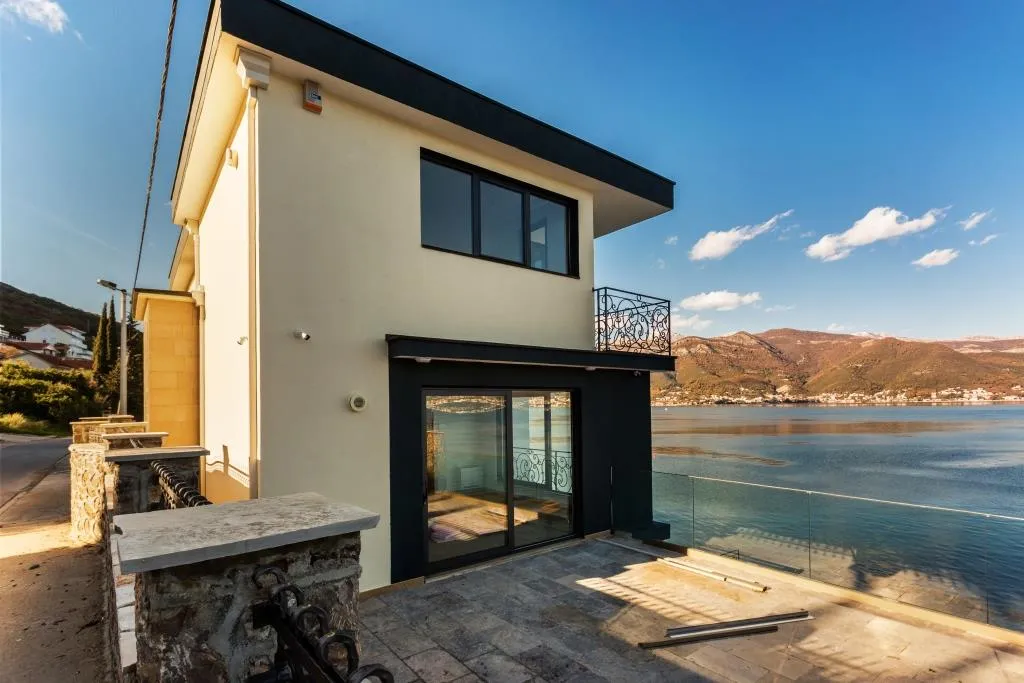 Exterior view of a modern two-story waterfront house featuring a stone terrace and large glass sliding doors.