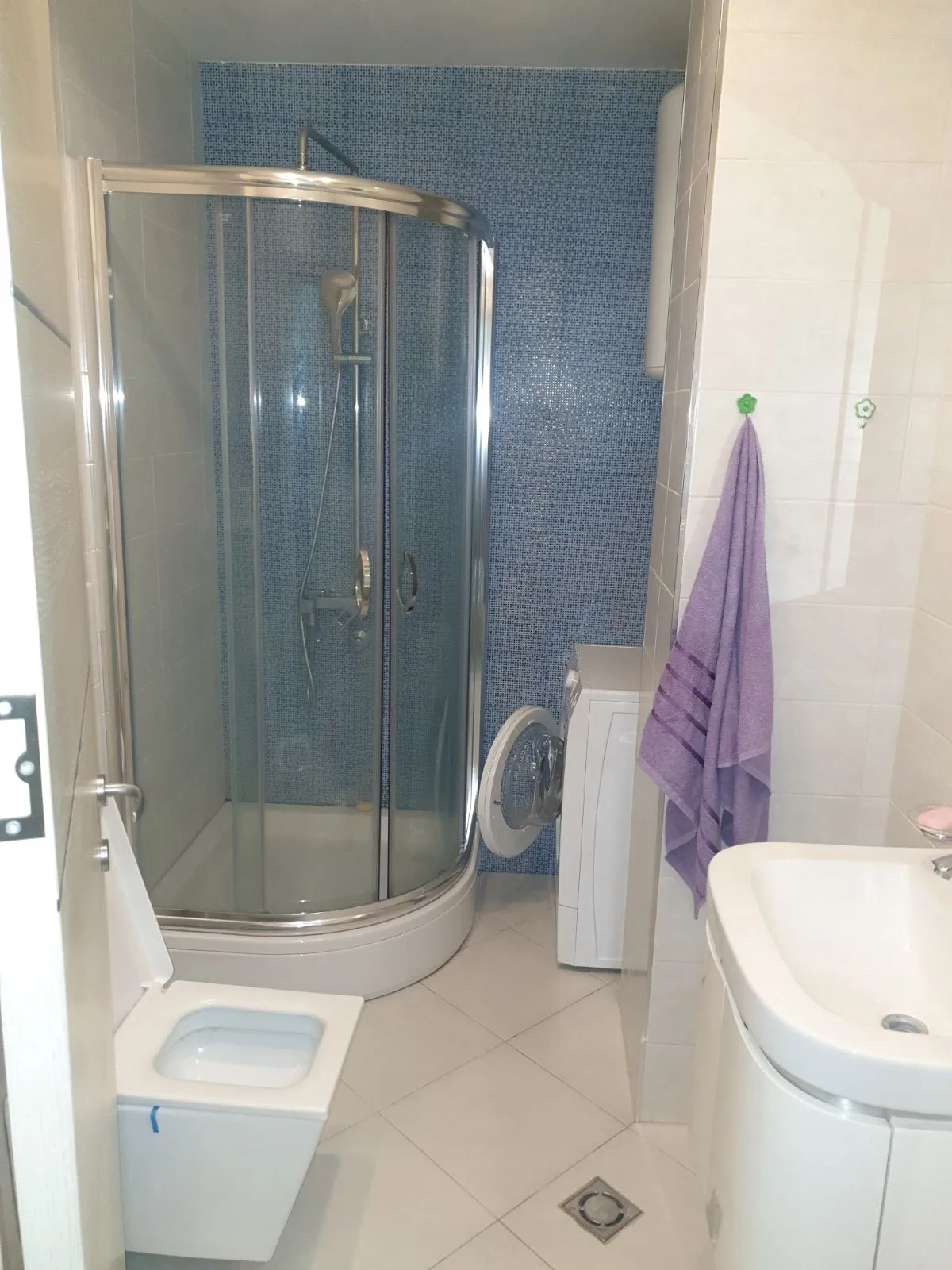 Bathroom featuring a glass shower enclosure, toilet, sink, and washing machine against blue mosaic tiles.