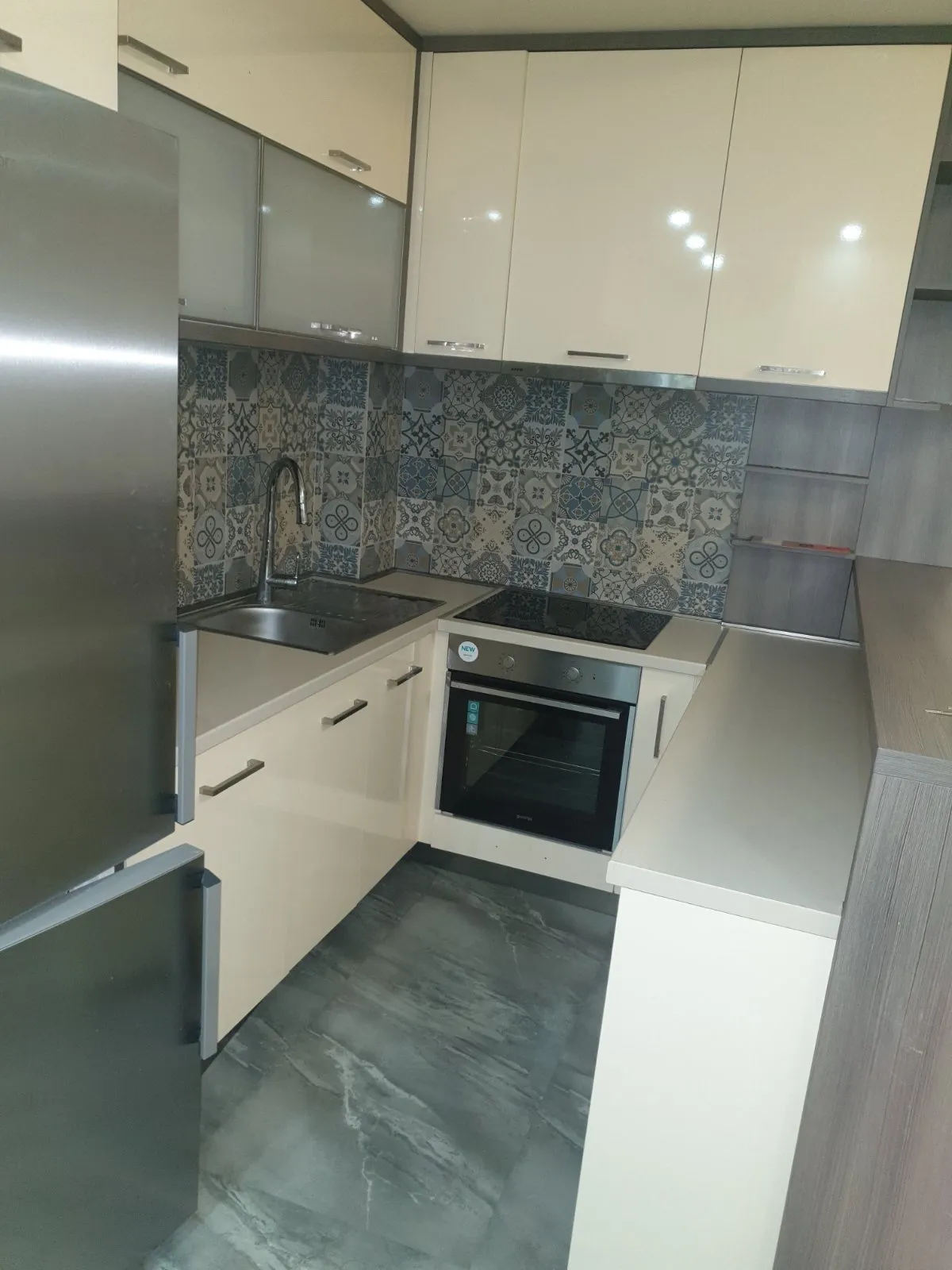 Modern kitchen featuring white cabinets, patterned wall tiles, built-in oven, cooktop, stainless steel sink, and refrigerator.