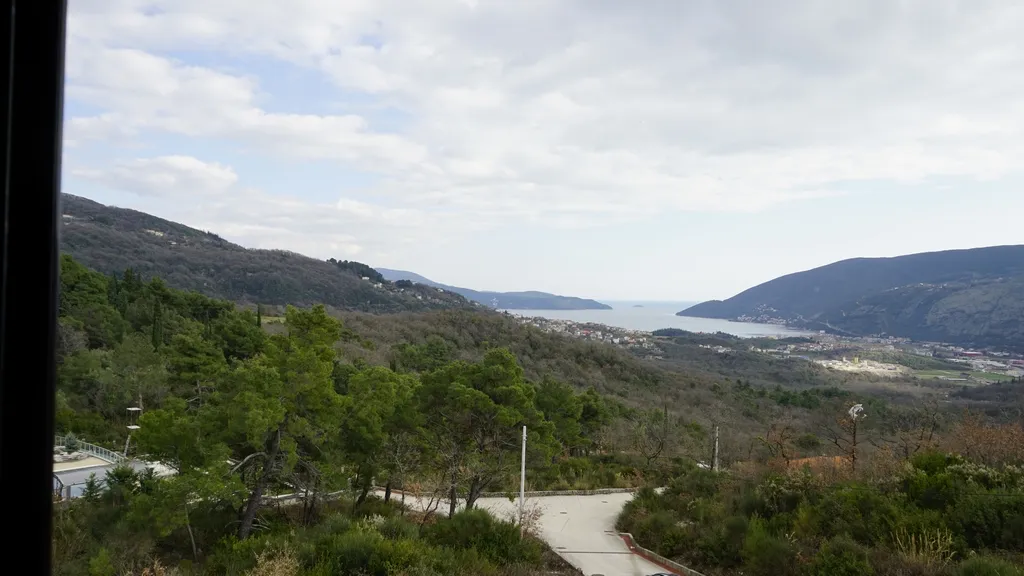 Panoramic view of hills, forest, and sea bay from a road. Black window frame visible on the left edge.