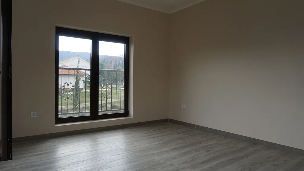 Empty room with grey laminate flooring and beige walls. Large window with dark frame and metal security bars.
