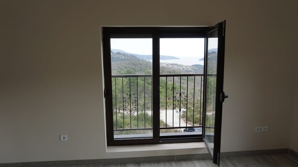 Sea and mountain view from the villa living room in Igalo, Montenegro: panoramic window with wrought-iron railing and light finishes.