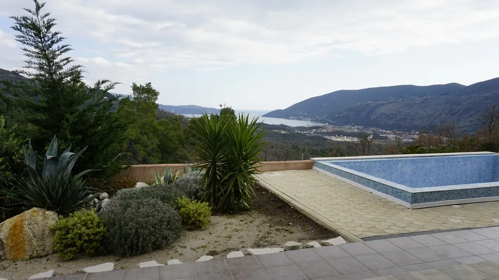 Blue tiled pool on a patio surrounded by plants with a view of mountains and sea.