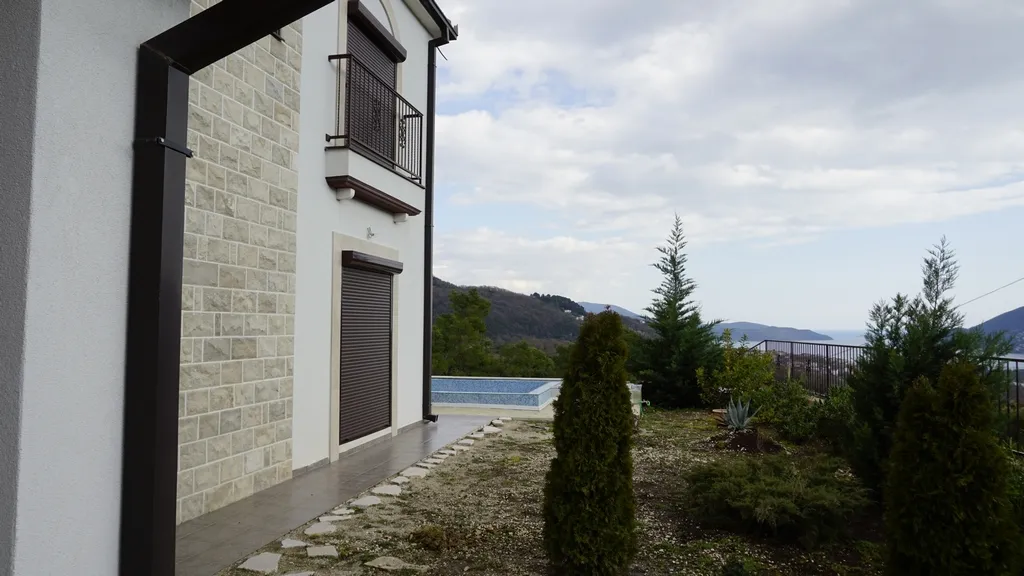 View of the house facade with stone cladding and terrace. A pool edge and mountain landscape are visible in the background garden.
